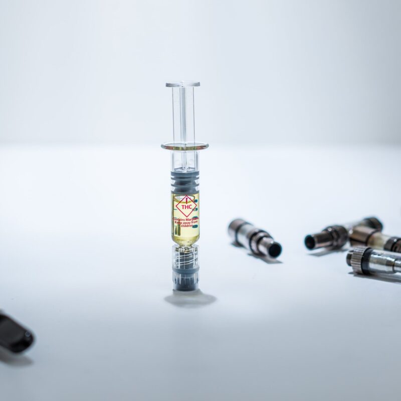 A THC distillate syringe standing upright, in the middle of used vape cartridges, and a vape pen on the other side. Spot light, white surface and background with dramatic light.